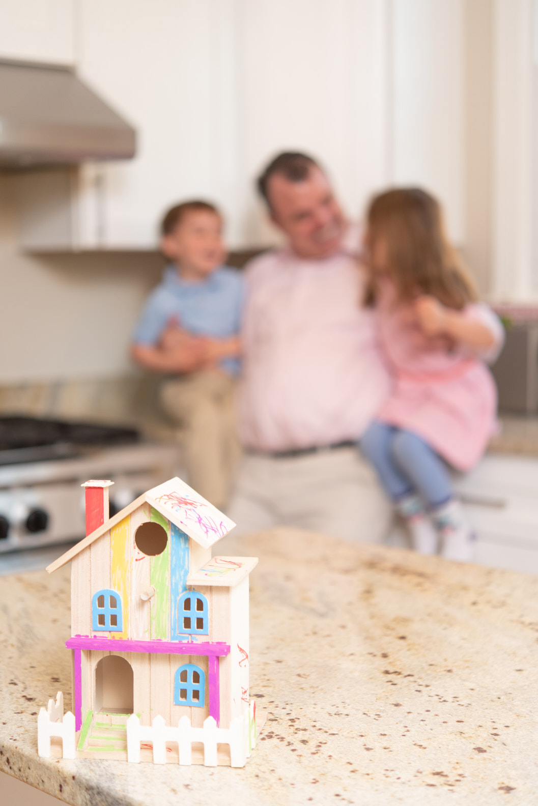 A father with two children, a colored birdhouse on the kitchen counter.